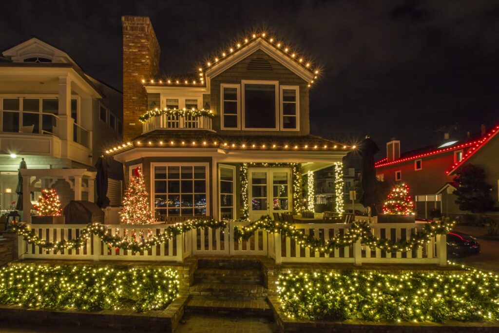 Festive beach house at night, decorated with bright Christmas lights along the roofline and fence for a dazzling holiday display. Garlands and small illuminated trees add to the cheerful scene.