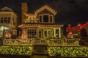 Festive beach house at night, decorated with bright Christmas lights along the roofline and fence for a dazzling holiday display. Garlands and small illuminated trees add to the cheerful scene.