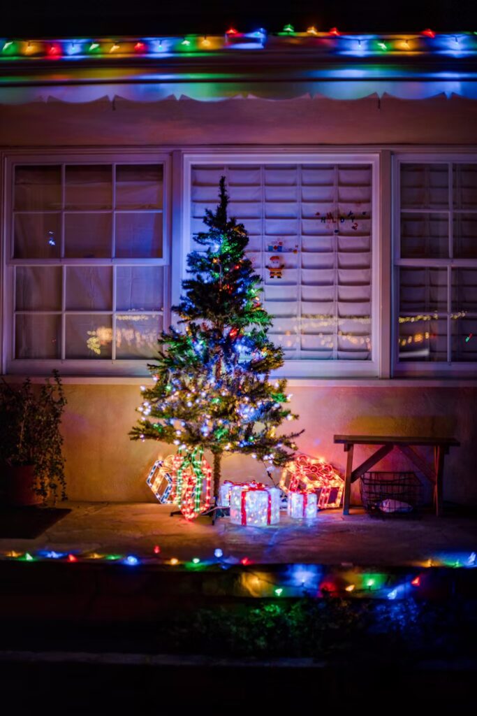 A small Christmas tree sparkles with lights on a porch, surrounded by glowing gift boxes. Colorful Christmas lights frame the roofline and porch edge, creating a festive holiday scene.