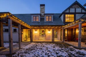 Exterior of a charming home decorated for Christmas with string lights, a wreath, and small Christmas trees. Snow dusts the ground in the courtyard, creating a magical holiday display.