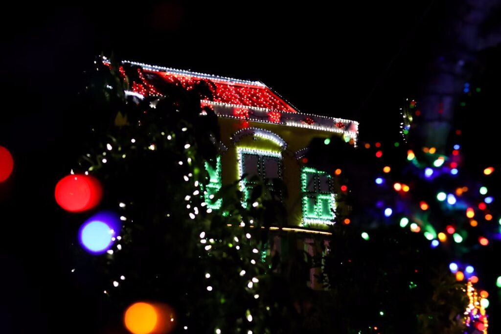 House adorned with outdoor Christmas lights, featuring red lights on the roof and green lights around the windows. White lights twinkle in the foreground, creating a magical holiday display.