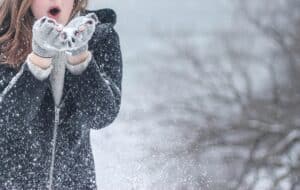 A woman in a dark winter coat and mittens blows snow from her hands.