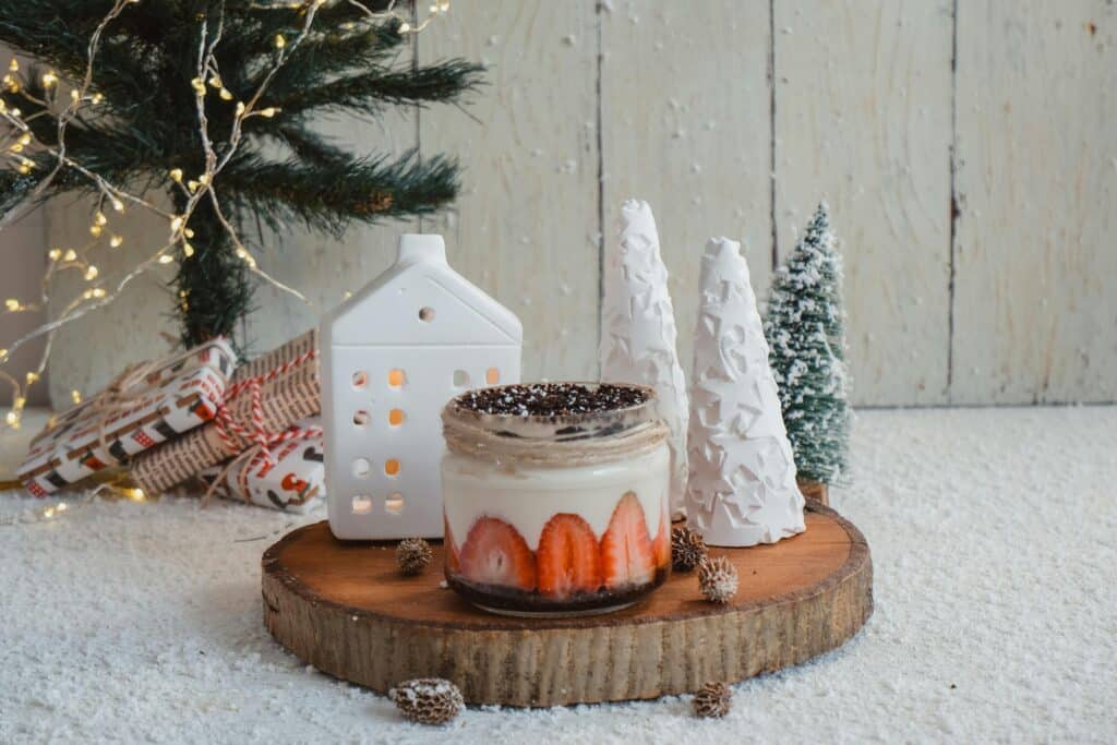 Festive Christmas dessert jar with strawberries and chocolate, set on a wooden slice with snowy decor, a mini Christmas tree, a ceramic house, and wrapped gifts. A cozy DIY Christmas decorations scene.