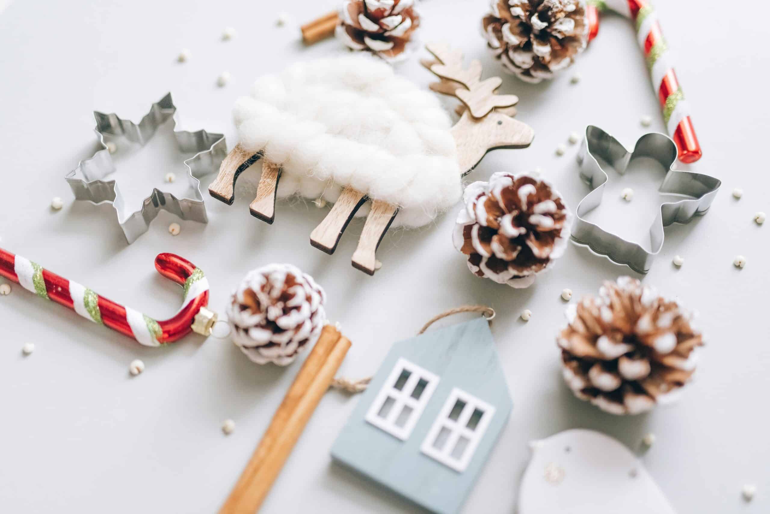 Festive DIY Christmas decoration elements: pine cones, candy canes, cookie cutters, a woolly sheep, and a wooden house ornament, arranged on a light gray surface.