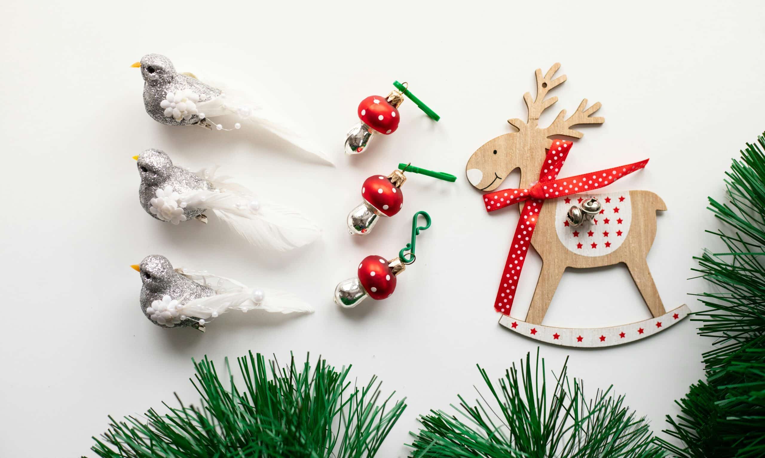 Festive DIY Christmas decorations: three glittery bird ornaments, red mushroom ornaments, a wooden reindeer with a red ribbon, and green pine sprigs on a white surface.