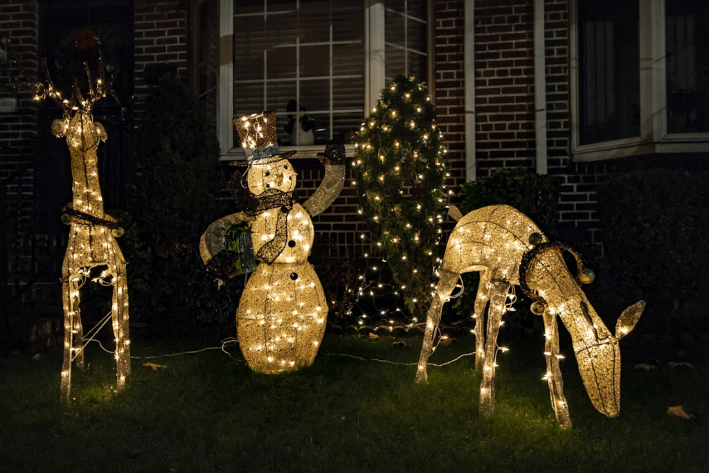 Glowing Christmas lights displays: Two wireframe reindeer and a snowman illuminate a lawn in front of a brick house. Holiday tradition transforming homes for Christmas.
