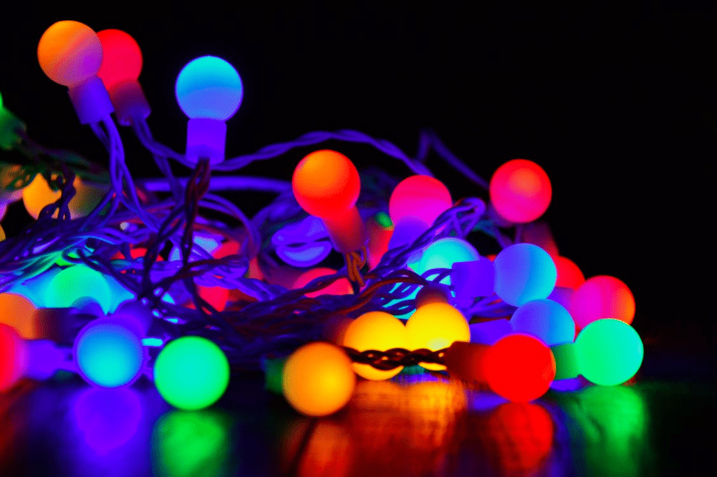 Colorful Christmas lights glow in a tangled pile, showing a close-up of the bulbs in shades of red, orange, blue, and green. The lights reflect on a dark surface, illustrating the challenge of storing Christmas lights without the tangle.