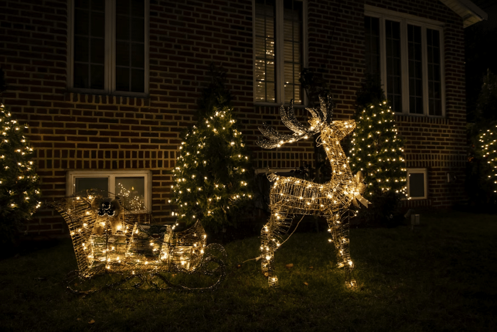 Illuminated Christmas lights display of a reindeer and sleigh on a lawn at night, with lit Christmas trees in the background against a brick house. A festive holiday tradition that transforms homes.