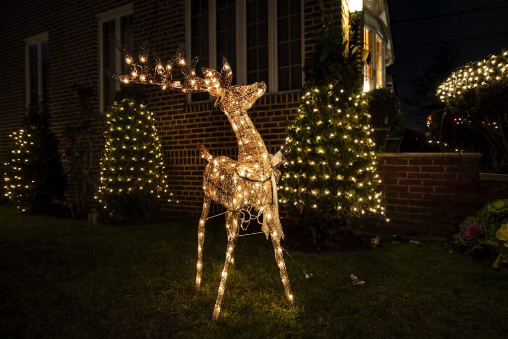 A lighted Christmas deer lawn decoration stands on green grass at night, part of a Christmas lights display in front of a brick house with lighted trees.