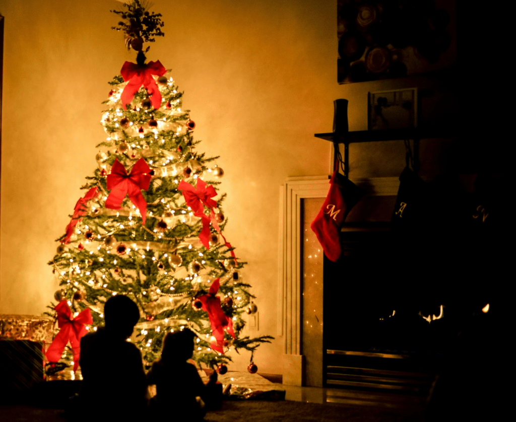 Warm Christmas scene: illuminated tree adorned with red bows and gold ornaments, casting a glow on a family silhouette. Stockings hang above a cozy, crackling fireplace.