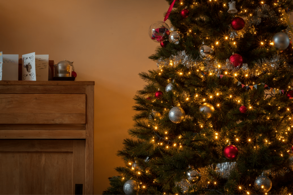 A warmly lit Christmas tree adorned with silver and red ornaments, and twinkling lights, stands next to a wooden cabinet with Christmas cards and a kettle on top, showcasing a festive holiday scene.