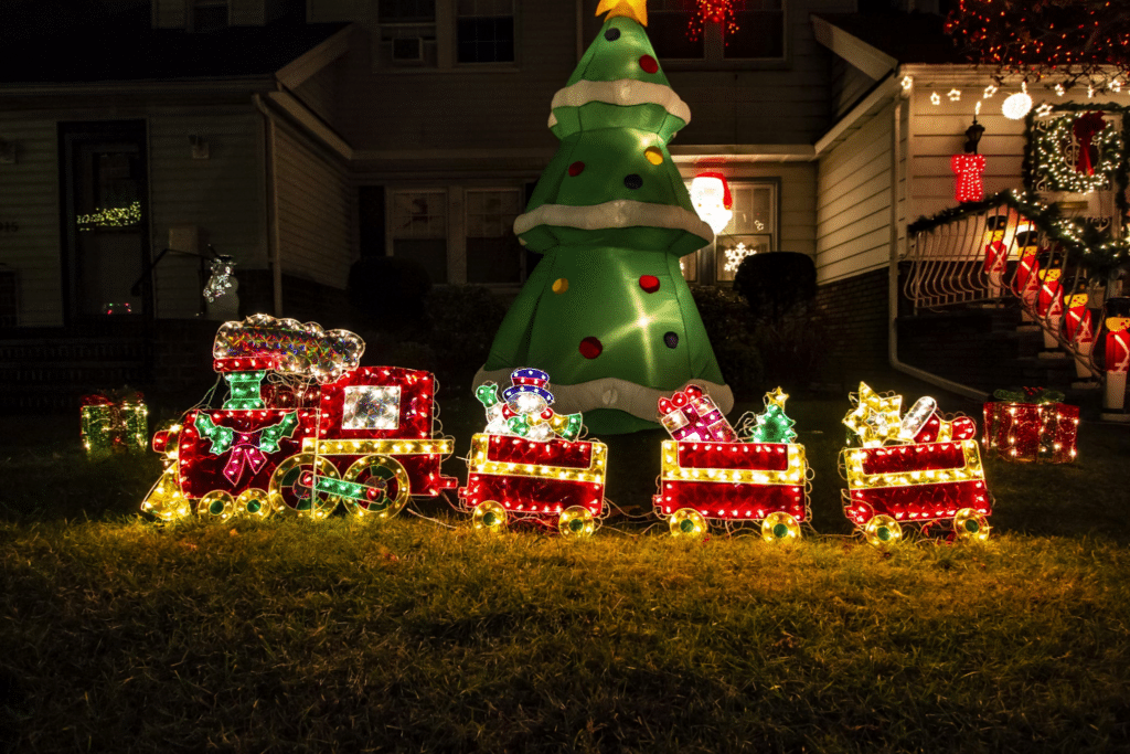Festive Christmas lights decorate a house, featuring an inflatable Christmas tree and a charming illuminated toy train on the lawn, adding holiday cheer to the neighborhood.