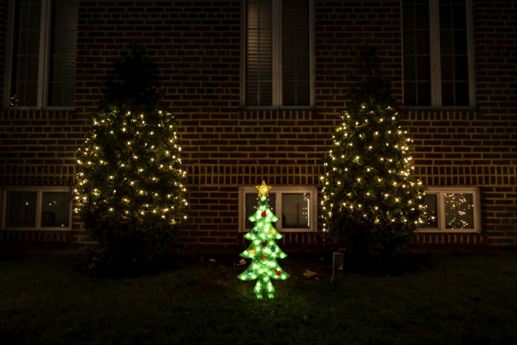 Warm white lights cover two shrubs beside an illuminated Christmas tree in front of a brick house.