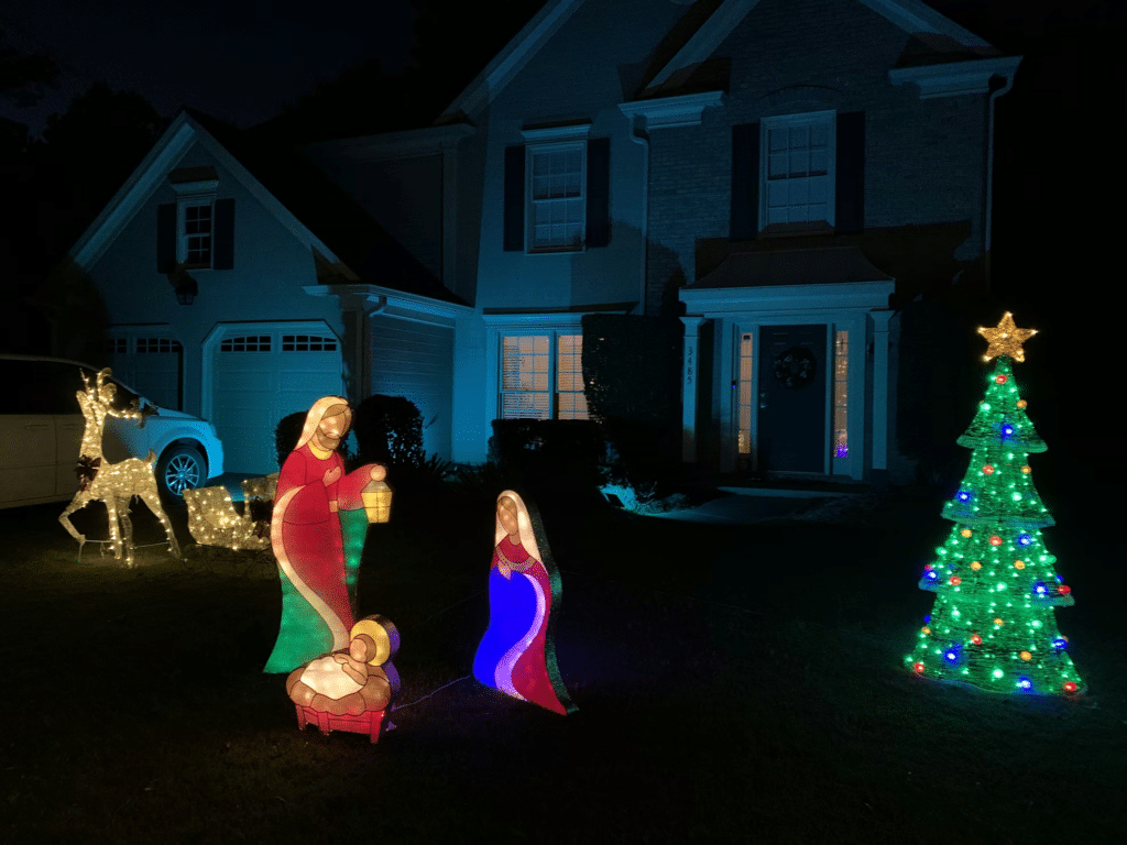 A lighted nativity scene, reindeer figures, and Christmas tree on the lawn in front of a house.