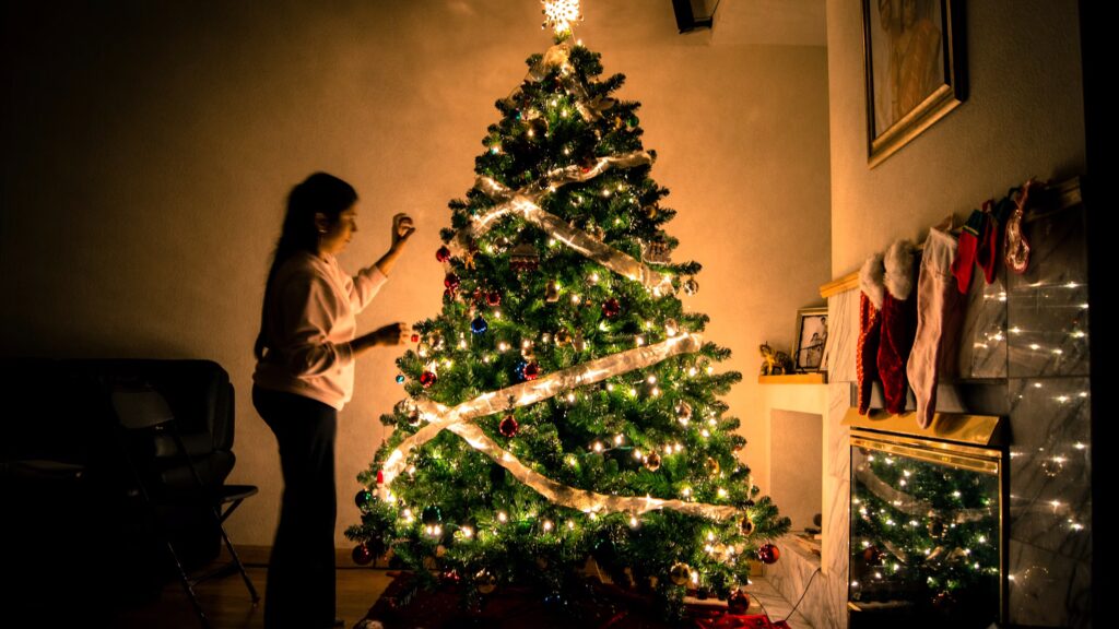 Woman decorating a Christmas tree with lights and ornaments in a warmly lit living room. The tree is adorned with a star on top and a ribbon, showcasing a step in "How to Put Lights on a Christmas Tree.