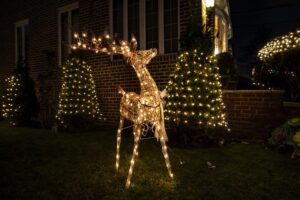 An illuminated wire reindeer stands on a lawn beside trees wrapped in white Christmas lights.