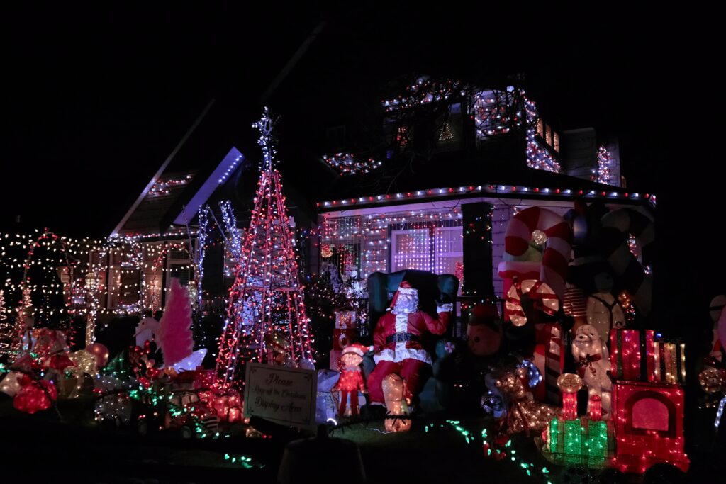 Extravagant Christmas lights adorn a house, featuring Santa in his chair, candy canes, a light-up tree, and a sign that reads "Area of Christmas Displaying Area." A festive and bright holiday scene.