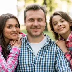 Three people smiling at camera outdoors: an adult in a checked shirt with two younger people in pink plaid leaning on the adult's shoulders.