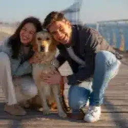 Two people crouch and smile while hugging a golden retriever on a wooden boardwalk by the water.